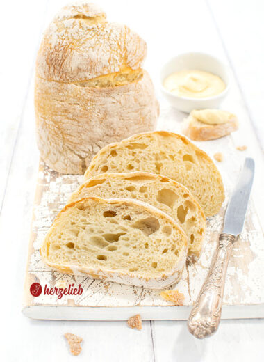 Drei Scheiben Pantoffelbrot vor einem auf der Schnittfläche stehenden Brotlaib. Seitlich neben dem Ciabatta ein Silbermesser, hinten ein Dip.