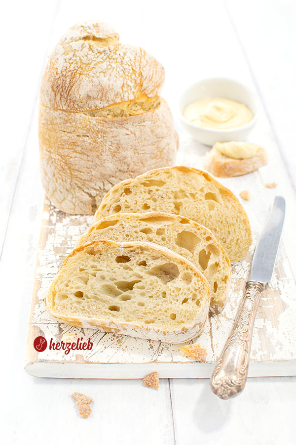 Drei Scheiben Pantoffelbrot vor einem auf der Schnittfläche stehenden Brotlaib. Seitlich neben dem Ciabatta ein Silbermesser, hinten ein Dip.