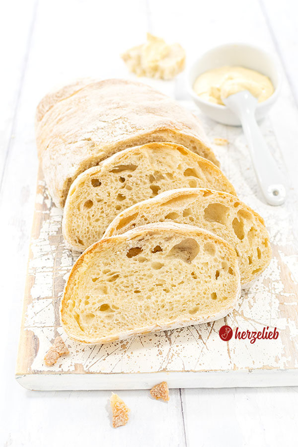 Einige Scheiben Pantoffelbrot vor einem angeschnittenen Brotlaib. Das Ciabatta liegt auf einem weißen Brett. Im Hintergrund ein Schälchen mit Dip.