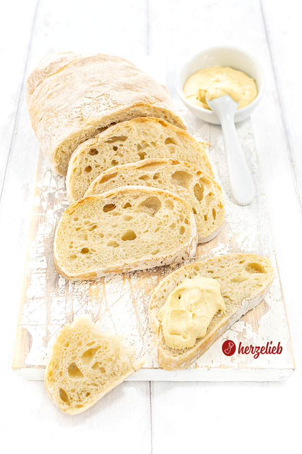 Luftiges Pantoffelbrot auf einem weißen Holzbrett. Im Vordergrund ein eine mit Dip bestrichene Scheibe Ciabatta. Im Hintergrund ein Dipschälchen.