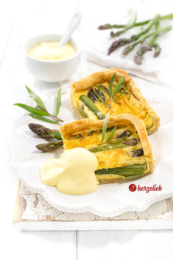 Zwei Stücke Spargeltarte mit Sauce Hollandaise auf einem Teller. Dekoriert mit Estragon und Spargelspitzen.