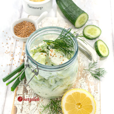 Creme-Schüttelgurken in einem Bügelglas. Dekoriert mit Dill und Senfsaat. Im Hintergrund Senf, Gurke und Senfsaat. Vorn eine halbe Zitrone und Dill.