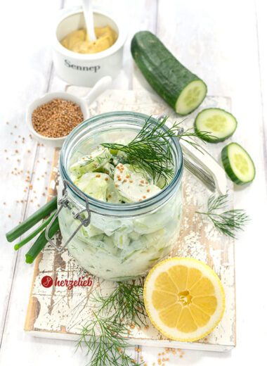 Creme-Schüttelgurken in einem Bügelglas. Dekoriert mit Dill und Senfsaat. Im Hintergrund Senf, Gurke und Senfsaat. Vorn eine halbe Zitrone und Dill.