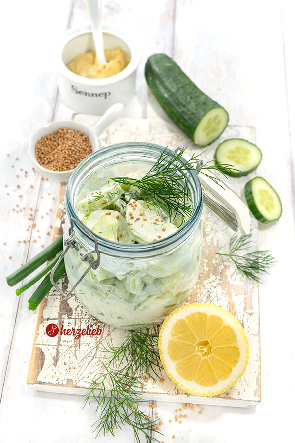Creme-Schüttelgurken in einem Bügelglas. Dekoriert mit Dill und Senfsaat. Im Hintergrund Senf, Gurke und Senfsaat. Vorn eine halbe Zitrone und Dill.