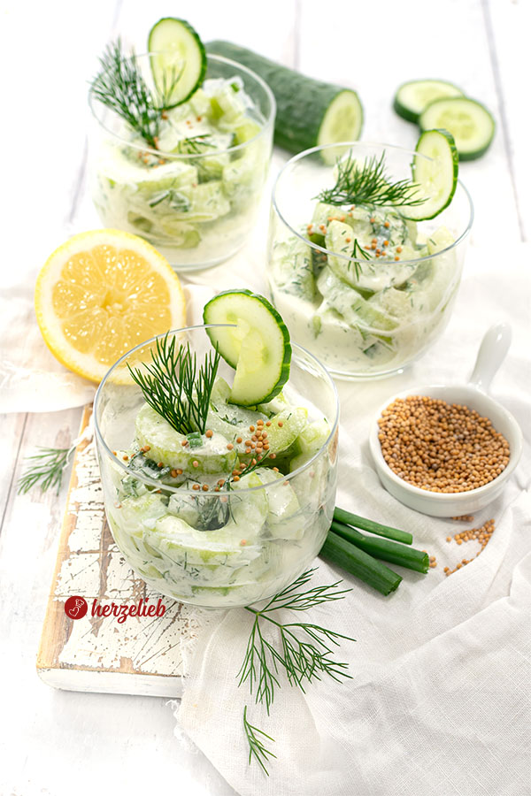 Creme-Schüttelgurken in drei Gläsern. Angerichtet und dekoriert mit Senfsaat und Dill. Hinten eine Zitrone und Gurke. Vorn Lauchzwiebeln und Senfsamen.