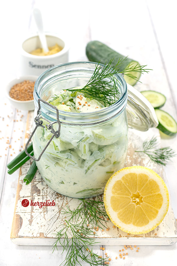Ein großes Bügelglas mit Creme-Schüttelgurken. Angerichtet auf einem Holzbrett, dekoriert mit Dill und Senfsaat. Im Vordergrund Zitrone, hinten Gurke.