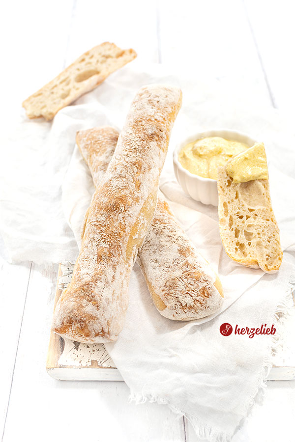 Links zwei Dinkelstangen, die übereinanderliegen. Rechts ein Schälchen Dip, an dem ein Stück Brot lehnt, das damit bestrichen ist.