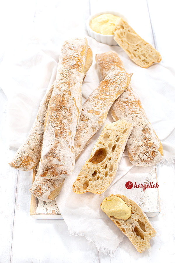 Übereinander liegen einige Dinkelstangen. Zwei aufgeschnittene Stücke von einer Brotstange liegen im Vordergrund, eines davon ist bestrichen mit Dip.
