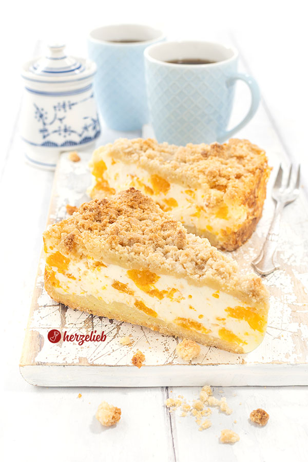 2 Stücke Mandarinen-Schmandkuchen auf einem Holzbrett. Rechts eine silberne Kuchengabel.Hinten 2 Becher Kaffeeund ein Zuckertopf