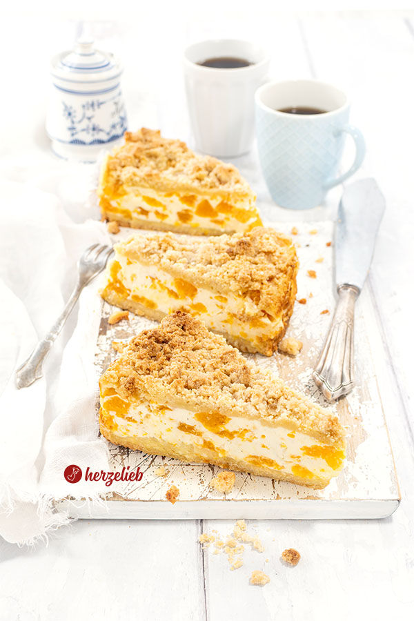 3 Stücke Mandarinen-Schmandkuchen wie von Oma auf einem Holzbrett. Rechts ein silbernes Tortenmesser. Hinten 2 Kaffeebecher und ein Zuckertopf