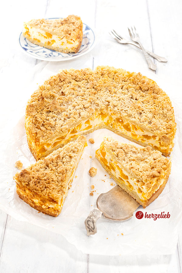 Ein angeschnittener Mandarinen-Schmandkuchen auf einem Stück Backpapier. 2 Stücke sind nach vorn gezogen, das rechte liegt auf einem Tortenheber.