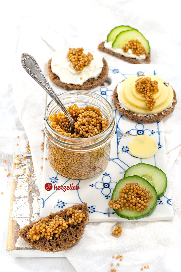 In der Mitte des Bildes ein Glas mit Senfkaviar mit einem Löffel darin. Rundherum Brot, Gurken, Käsebrot, Quarkbrot mit dem Kaviar.