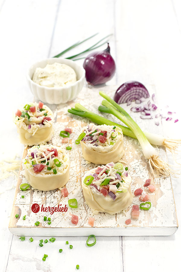 Vorbereitete, ungebacken Flammkuchen-Schnecken auf einem Holzbrett. Angerichtet mit Frühlingszwiebeln, roter Zwiebel und Frischkäse im Hintergrund.