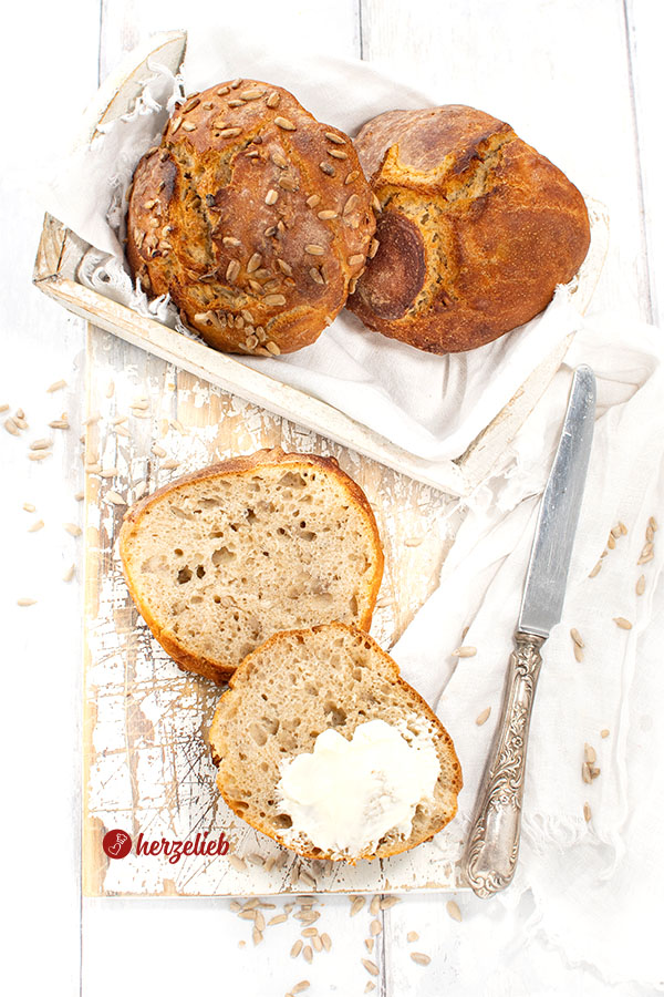 Zwei Sonnenblumkernbrötchen in einer Kiste, davor ein halbiertes Brötchen, eine Hälfte mit Butter bestrichen.