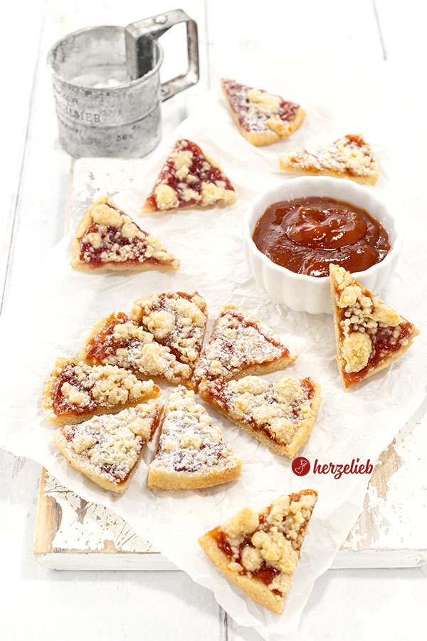 Im Vordergrund geschnittene Streuselkuchen-Kekse, mit Puderzucker bestäubt. Daneben weitere Plätzchen ohne Puderzuckerschicht. Rechts hinten eine Schale Marmelade.