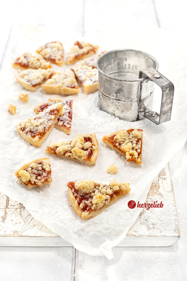 Schnelle Streuselkuchen-Kekse mit Marmelade, teilweise bestäubt mit Puderzucker. Rechts im Hintergrund ein silberfarbenes Puderzuckersieb.