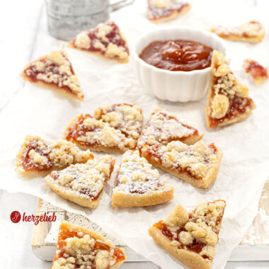 Streuselkuchen-Kekse mit Marmelade. Mit Puderzucker bestäubt. Im Hintergrund ein Schälchen Marmelade und ein Puderzuckersieb aus Metall.