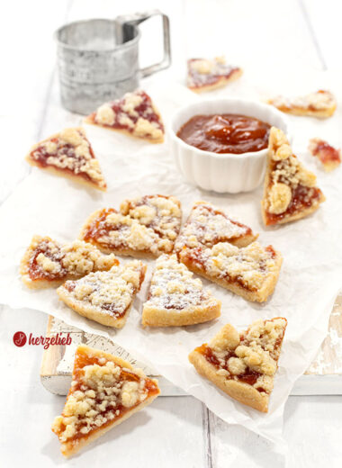 Streuselkuchen-Kekse mit Marmelade. Mit Puderzucker bestäubt. Im Hintergrund ein Schälchen Marmelade und ein Puderzuckersieb aus Metall.