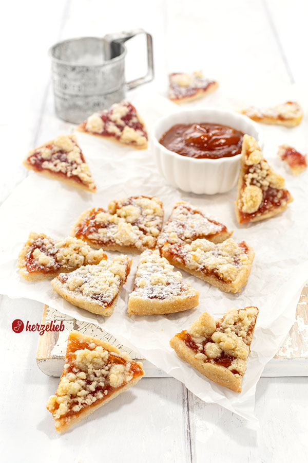 Streuselkuchen-Kekse mit Marmelade. Mit Puderzucker bestäubt. Im Hintergrund ein Schälchen Marmelade und ein Puderzuckersieb aus Metall. 