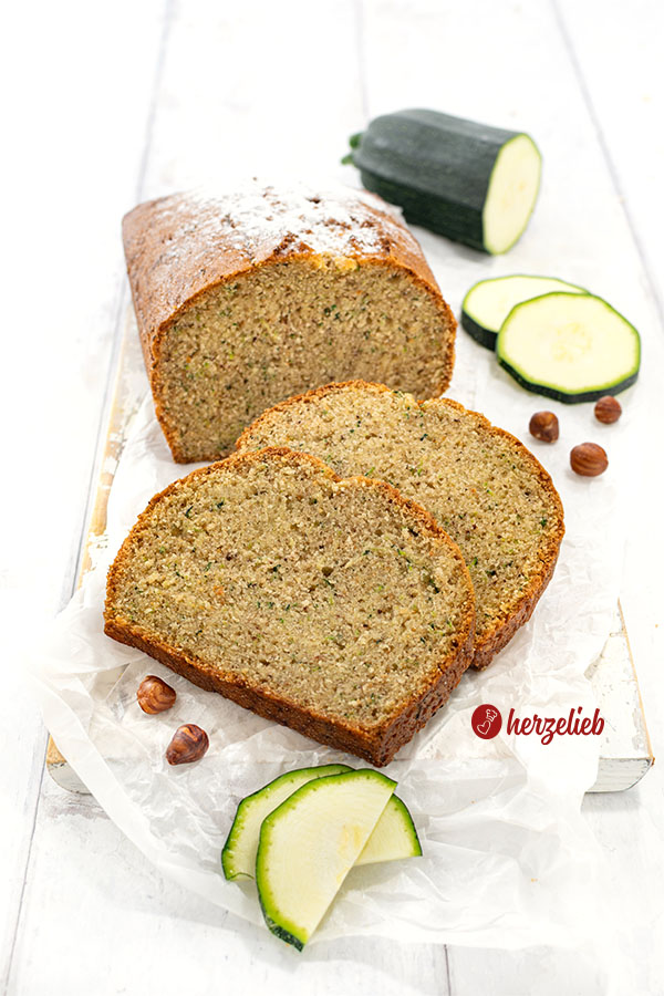 2 Scheiben Zucchinikuchen oder Nusskuchen vor einem angeschnittenen Kuchen auf einem Holzbrett. Dekoriert mit Haselnüssen und Zucchini.