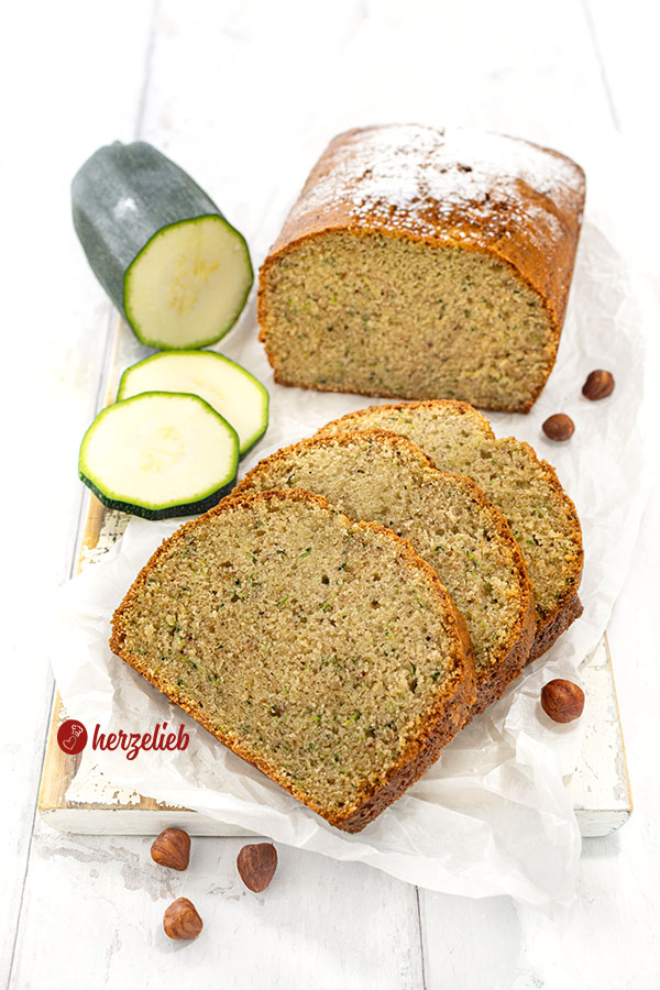 Nuss-Zucchinikuchen auf einem Brett. Vor dem angeschnittenen Nusskuchen liegen 3 Scheiben davon.