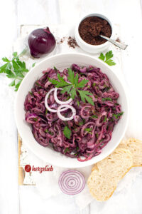Schale türkischer Zwiebelsalat. Dekoriert mit glatter Petersilie und Zwiebelringen. Im Hintergrund Sumach und eine rote Zwiebel