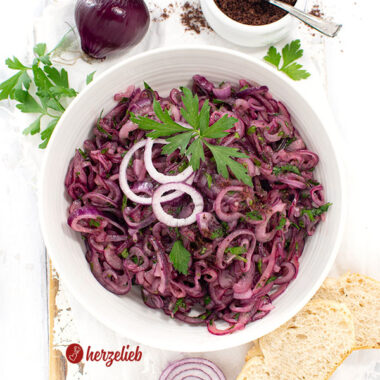 Schale türkischer Zwiebelsalat. Dekoriert mit glatter Petersilie und Zwiebelringen. Im Hintergrund Sumach und eine rote Zwiebel