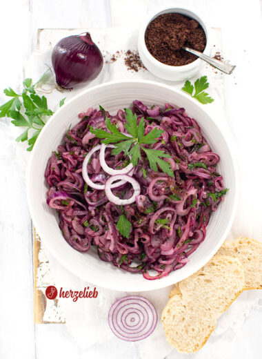Schale türkischer Zwiebelsalat. Dekoriert mit glatter Petersilie und Zwiebelringen. Im Hintergrund Sumach und eine rote Zwiebel
