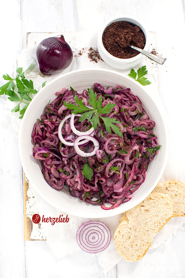 Schale türkischer Zwiebelsalat. Dekoriert mit glatter Petersilie und Zwiebelringen. Im Hintergrund Sumach und eine rote Zwiebel
