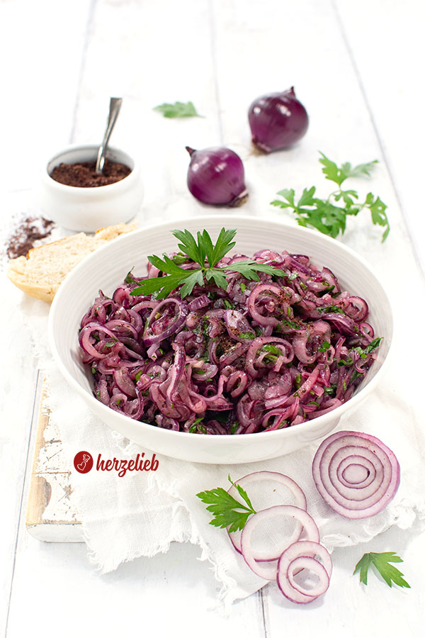 Würziger türkischer Zwiebelsalat in einer Schüssel. Dekoriert mit glatter Petersilie. Im Hintergrund rote Zwieeln, Sumach und Petersilie
