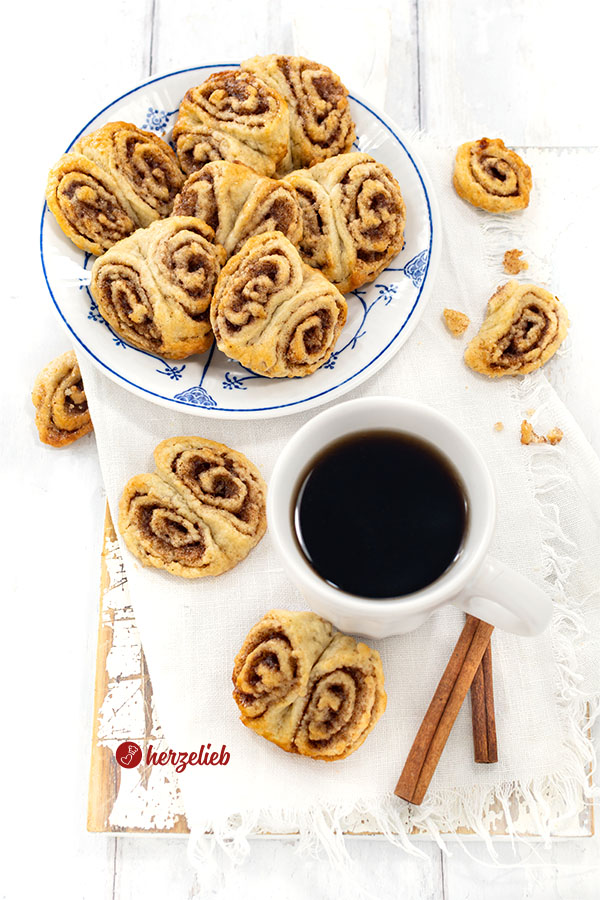 Franzbrötchen-Kekse auf einem Teller und eine Tasse Kaffee, um die herum weitere Kekse lieben