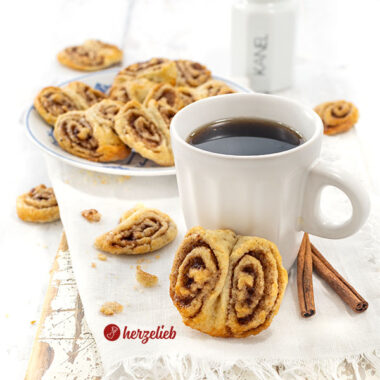 Eine Tasse Kaffee an einer der Franzbrötchen-Kekse, das nach dem Plätzchen-Rezept von herzelieb gebacken ist. Im Hintergrund weitere Kekse.