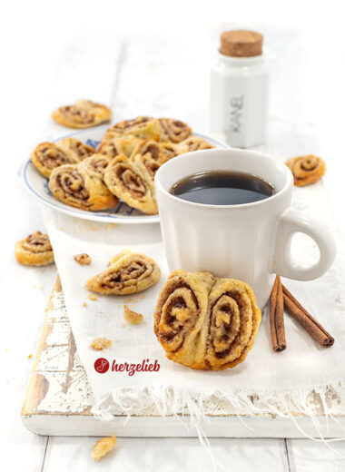 Eine Tasse Kaffee an einer der Franzbrötchen-Kekse, das nach dem Plätzchen-Rezept von herzelieb gebacken ist. Im Hintergrund weitere Kekse.