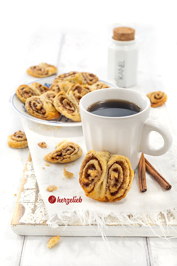 Eine Tasse Kaffee an einer der Franzbrötchen-Kekse, das nach dem Plätzchen-Rezept von herzelieb gebacken ist. Im Hintergrund weitere Kekse.