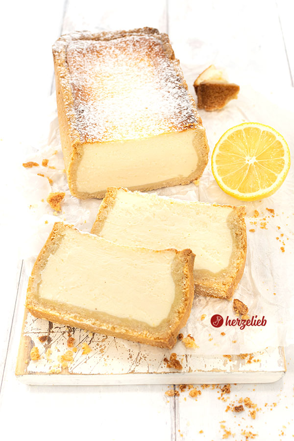 Käsekuchen aus der Kastenform auf einem Holzbrett. Vor dem angeschnittenen Kuchen liegen zwei Scheiben davon.