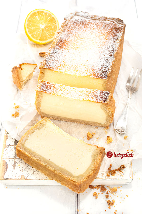 Goldbraun gebackener Käsekuchen aus der Kastenform. Angeschnitten, mit zwei Scheiben vor dem angeschnittenen Käsekuchen