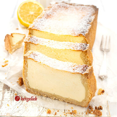 Saftiger Käsekuchen aus der Kastenform. Der angeschnittene Kuchen steht auf einem Holzbrett, davor stehen aufrecht zwei Scheiben davon.