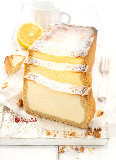 Saftiger Käsekuchen aus der Kastenform. Der angeschnittene Kuchen steht auf einem Holzbrett, davor stehen aufrecht zwei Scheiben davon.