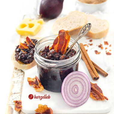 Dunkle Speckmarmelade nach einem Bacon Jam Rezept von herzelieb. Dekoriert mit knusprigen Speckstücken und einer Zwiebel. Angerichtet mit Brot und Käse.
