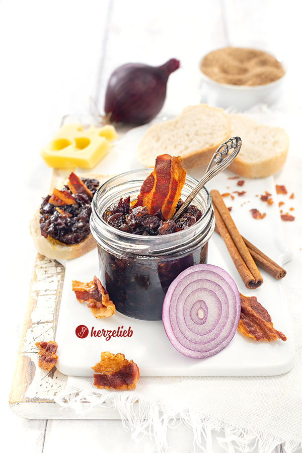 Dunkle Speckmarmelade nach einem Bacon Jam Rezept von herzelieb. Dekoriert mit knusprigen Speckstücken und einer Zwiebel. Angerichtet mit Brot und Käse.