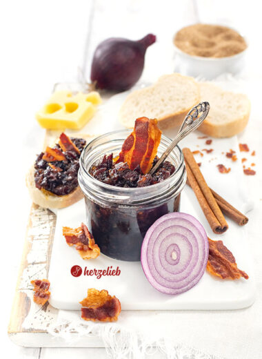 Dunkle Speckmarmelade nach einem Bacon Jam Rezept von herzelieb. Dekoriert mit knusprigen Speckstücken und einer Zwiebel. Angerichtet mit Brot und Käse.