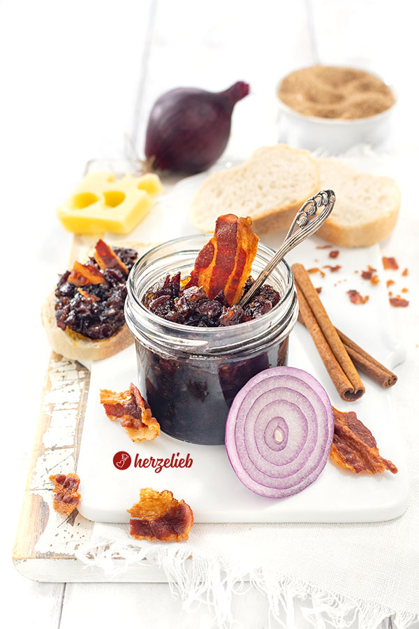Dunkle Speckmarmelade nach einem Bacon Jam Rezept von herzelieb. Dekoriert mit knusprigen Speckstücken und einer Zwiebel. Angerichtet mit Brot und Käse.