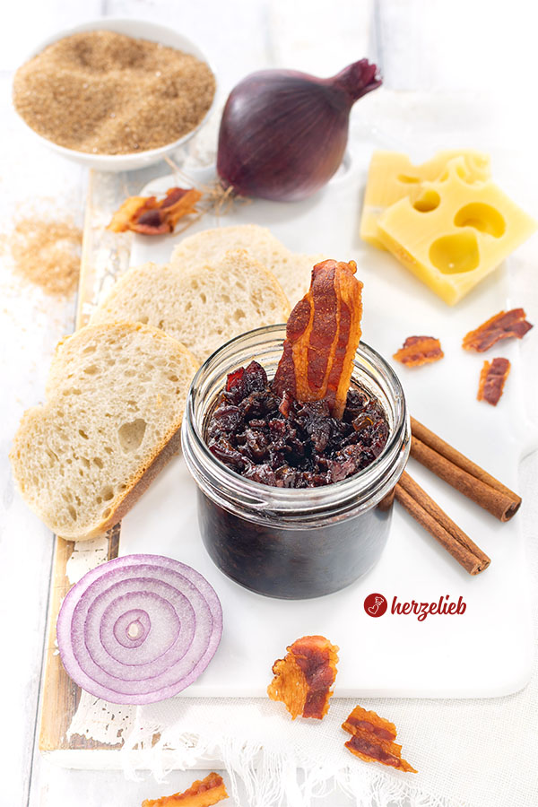 Ein Glas Speckmarmelade nach einem Bacon Jam Rezept von herzelieb. Dekoriert mit knusprigem Speck, Brot, Käse und Zimstangen.