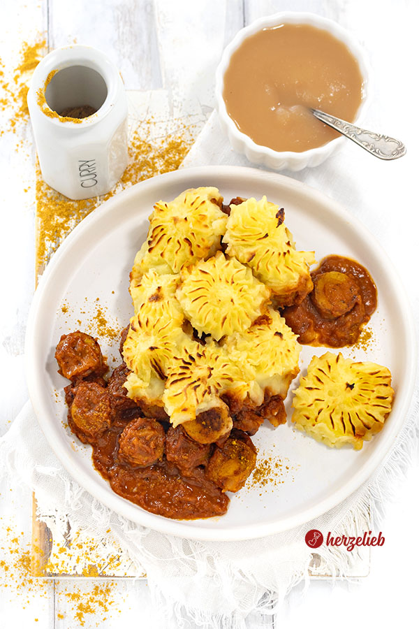 Teller mit Currywurstauflauf mit Kartoffelhaube. Dekoriert mit Currypulver, Apfelmus und Gewürzstreuern.