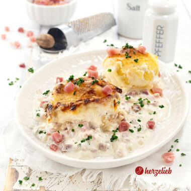 Ein Teller mit 2 mit Käse überbackenen Kartoffeln nach einem Flammkuchen-Kartoffeln Rezept von herzelieb. Angerichtet mit viel heller Soße.