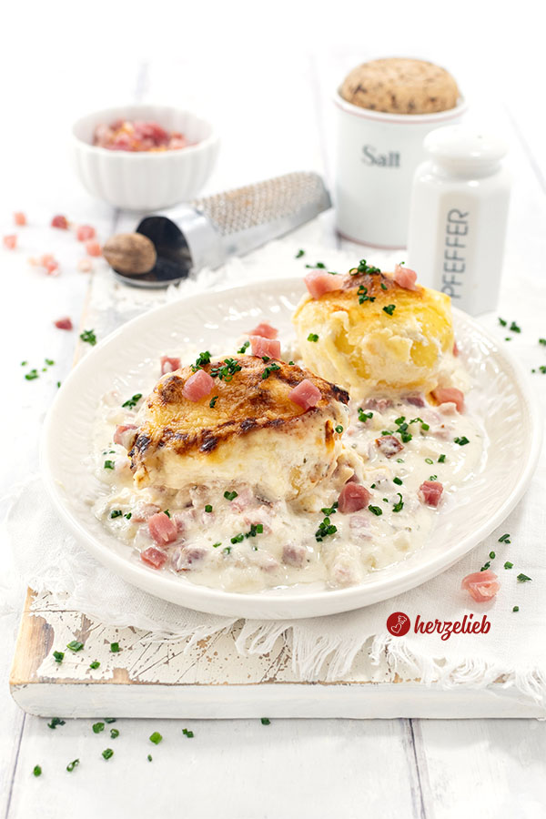 Ein Teller mit 2 mit Käse überbackenen Kartoffeln nach einem Flammkuchen-Kartoffeln Rezept von herzelieb. Angerichtet mit viel heller Soße.