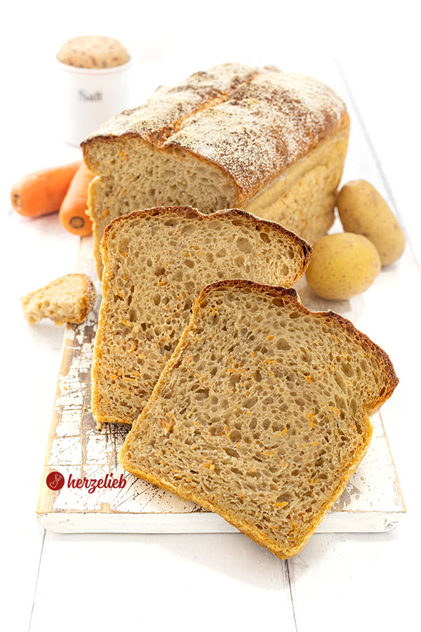 Angeschnittenes Kartoffel-Karotte-Brot auf ienem Holzbrett. Vor dem Brotlaib stehen zwei Scheiben davon. 