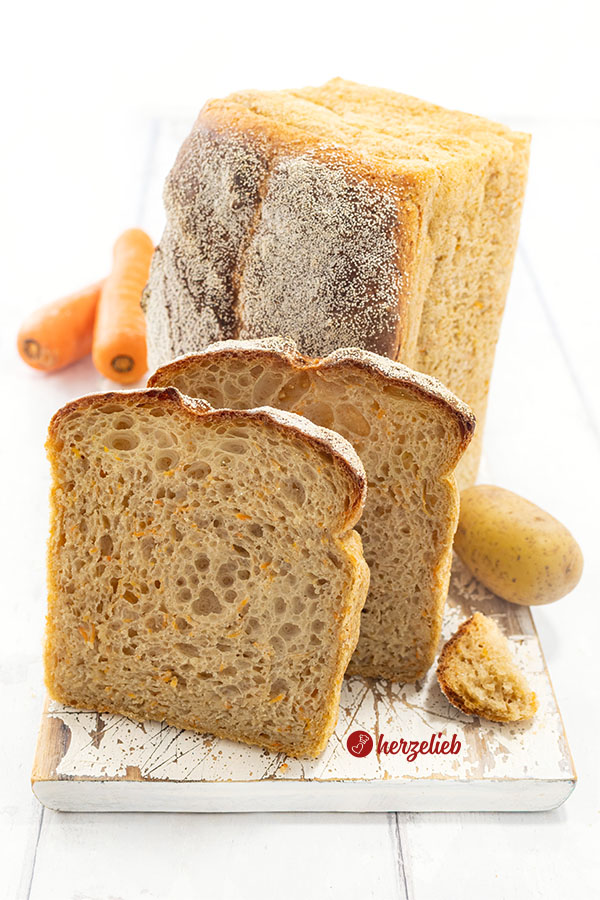 Leckeres Kartoffel-Karotten-Brot auf einem Holzbrett. Im Hintergrund steht auf der Schnittfläche der Brotlaib, davor stehen zwei Scheiben davon.