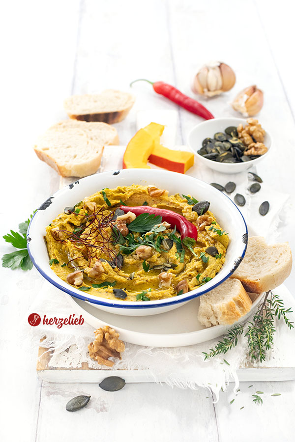 Eine Emailleschüssel mtit Ofenkürbis-Dip mit Feta. Serviert mit Brot, dekoriert mit Walnüssen, Kürbiskernen, Chilifäden, Petersilie und roter Chilischote.