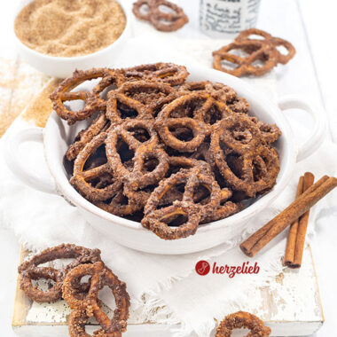Eine Schüssel mit Knabberkram nach einem Rezept für gebrannte Salzbrezeln von herzelieb. Dekoriert mit Zimtstangen. Im Hintergrund Gewürzgefäße